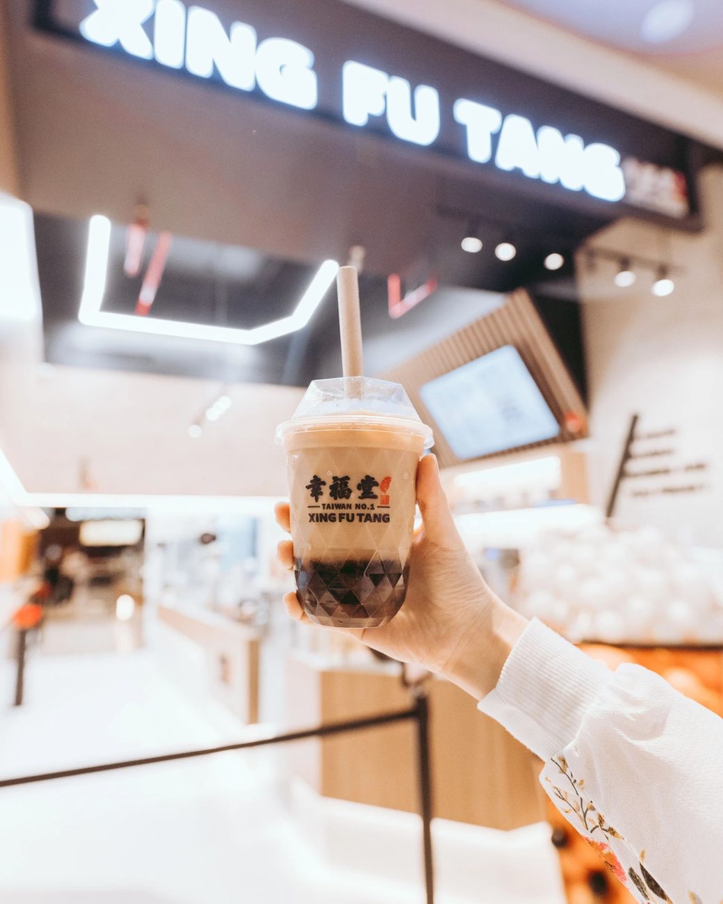 Mano sosteniendo un vaso de boba milk de Xing Fu Tang frente al letrero iluminado de la tienda