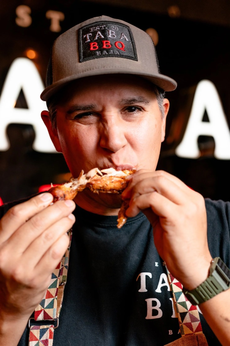 El chef Kris Kabush, fundador de Taba BBQ, mordiendo una alita frente al letrero de la marca