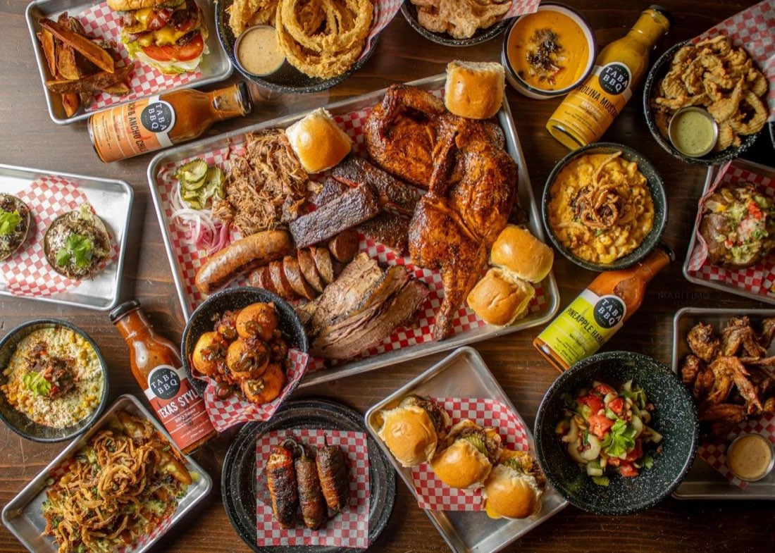 Vista aérea de una mesa de Taba BBQ con charolas de carnes ahumadas, sliders, alitas, guarniciones y salsas