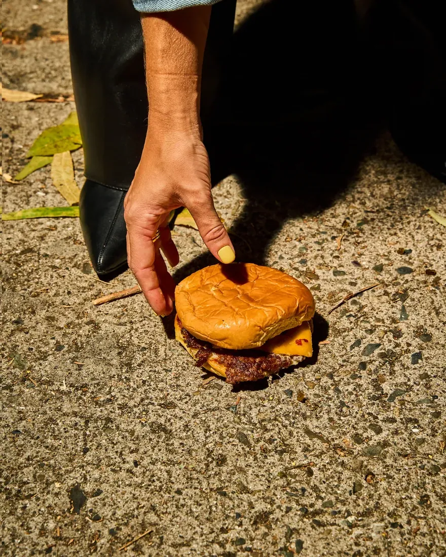 Mano con uñas amarillas recogiendo una smash burger de Naked Lukas en la acera