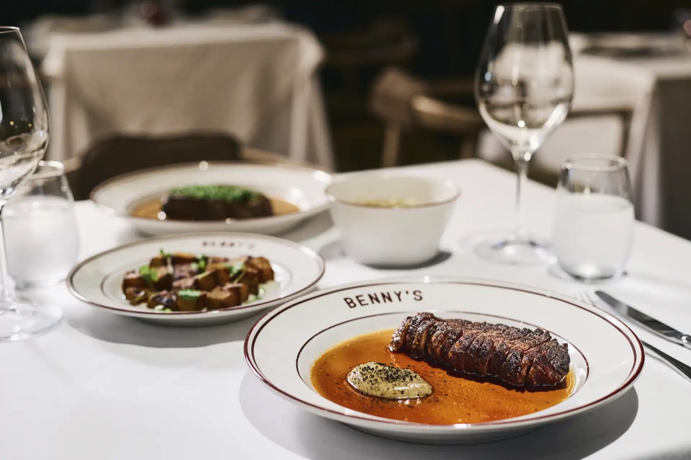 Mesa de Benny's con un corte de carne en salsa, copas de vino y platos de la casa