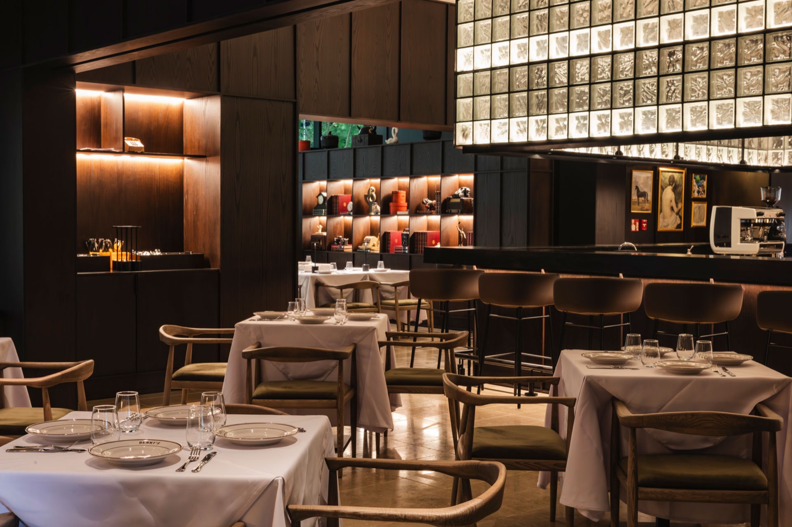 Salón de Benny's con manteles blancos, sillas de madera y la barra iluminada al fondo