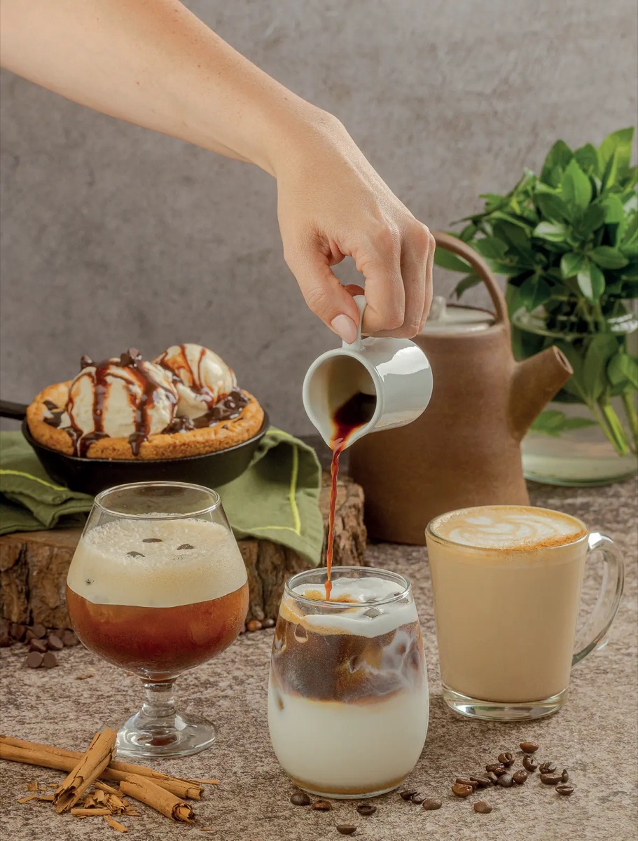 Barista sirviendo espresso sobre leche fría junto a un latte caliente y un cookie skillet en Tagers