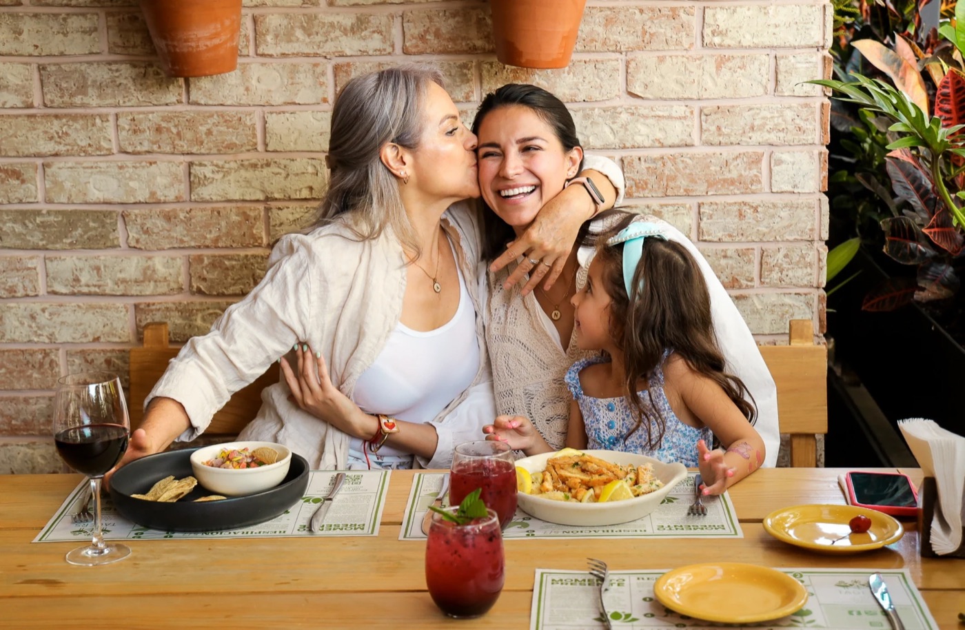 Abuela, mamá e hija abrazándose y riendo en una mesa de Tagers con platillos y bebidas