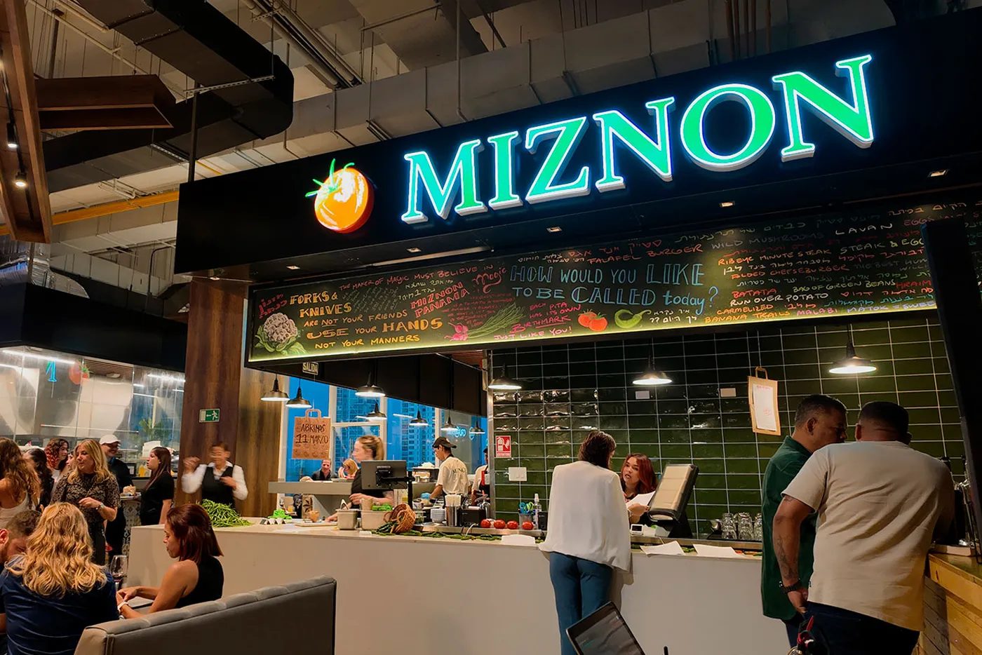 Local de Miznon en Pacific Center, Panamá, con su letrero de neón, pared de azulejos verdes y clientes en la barra