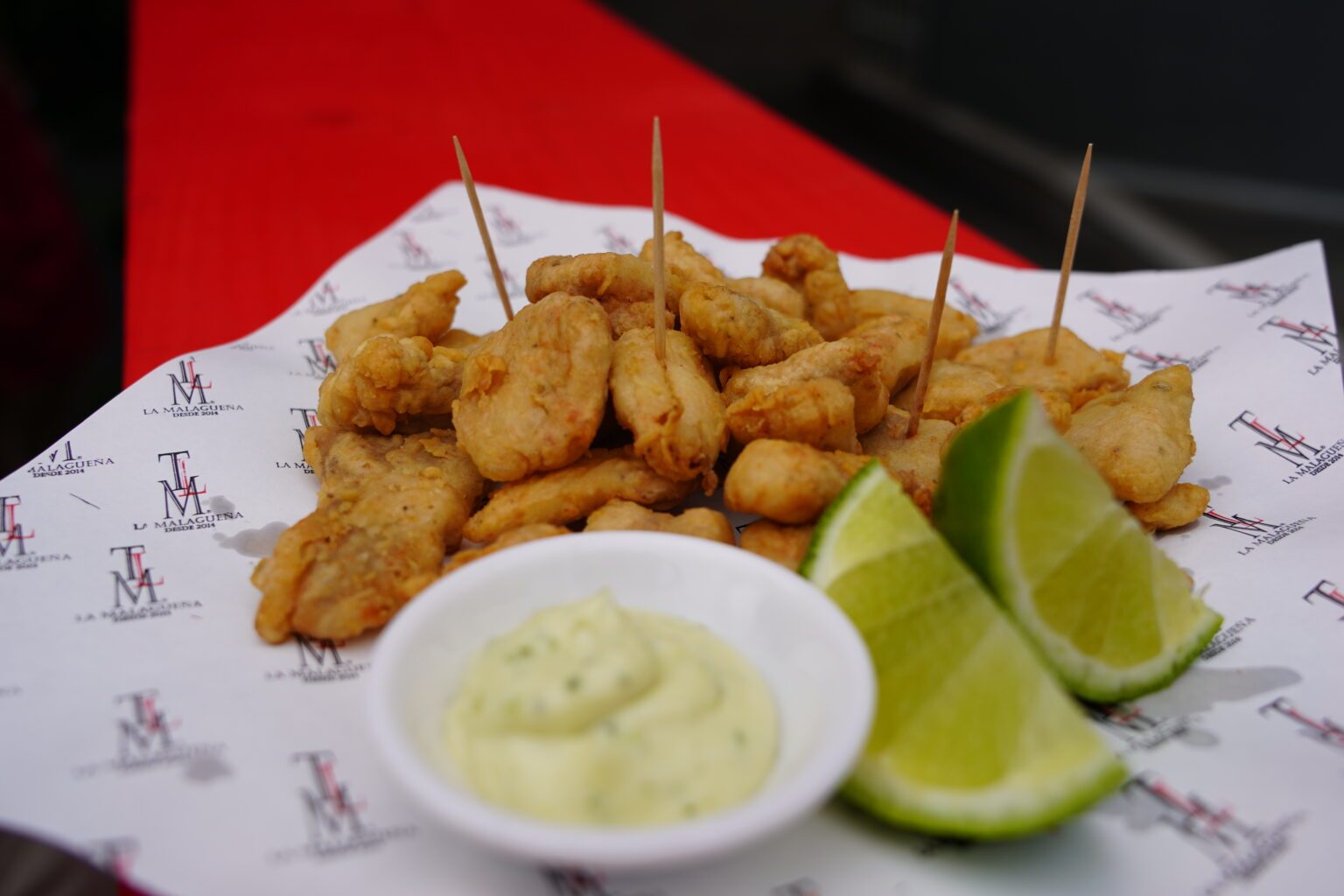 Frituras andaluzas doradas con alioli y limón sobre papel con el logo de La Malagueña