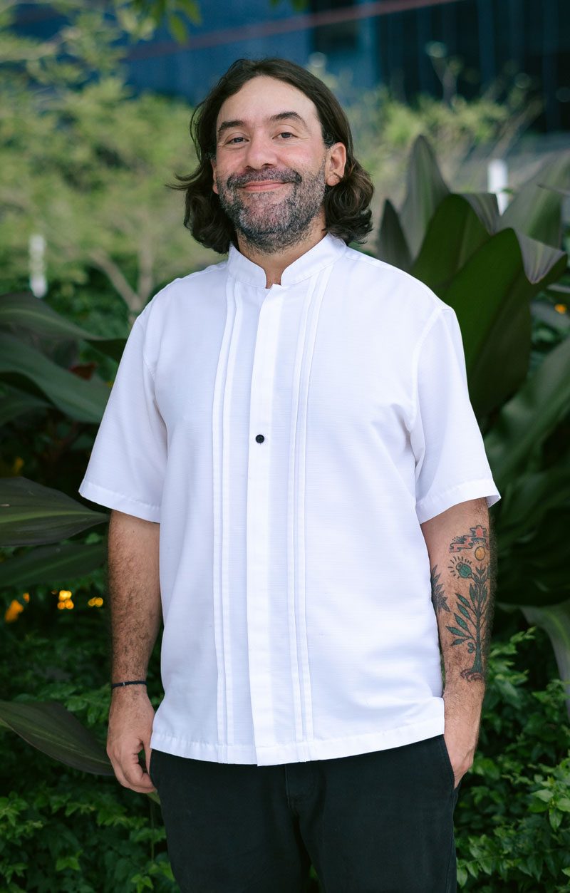 El chef Mario Castrellón sonriendo con filipina blanca entre plantas tropicales